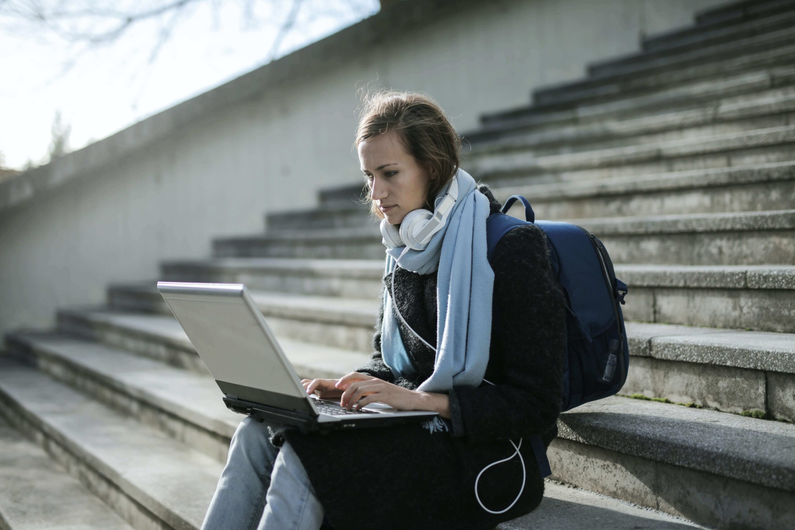 A woman sat on outside stairs while typing on a laptop.