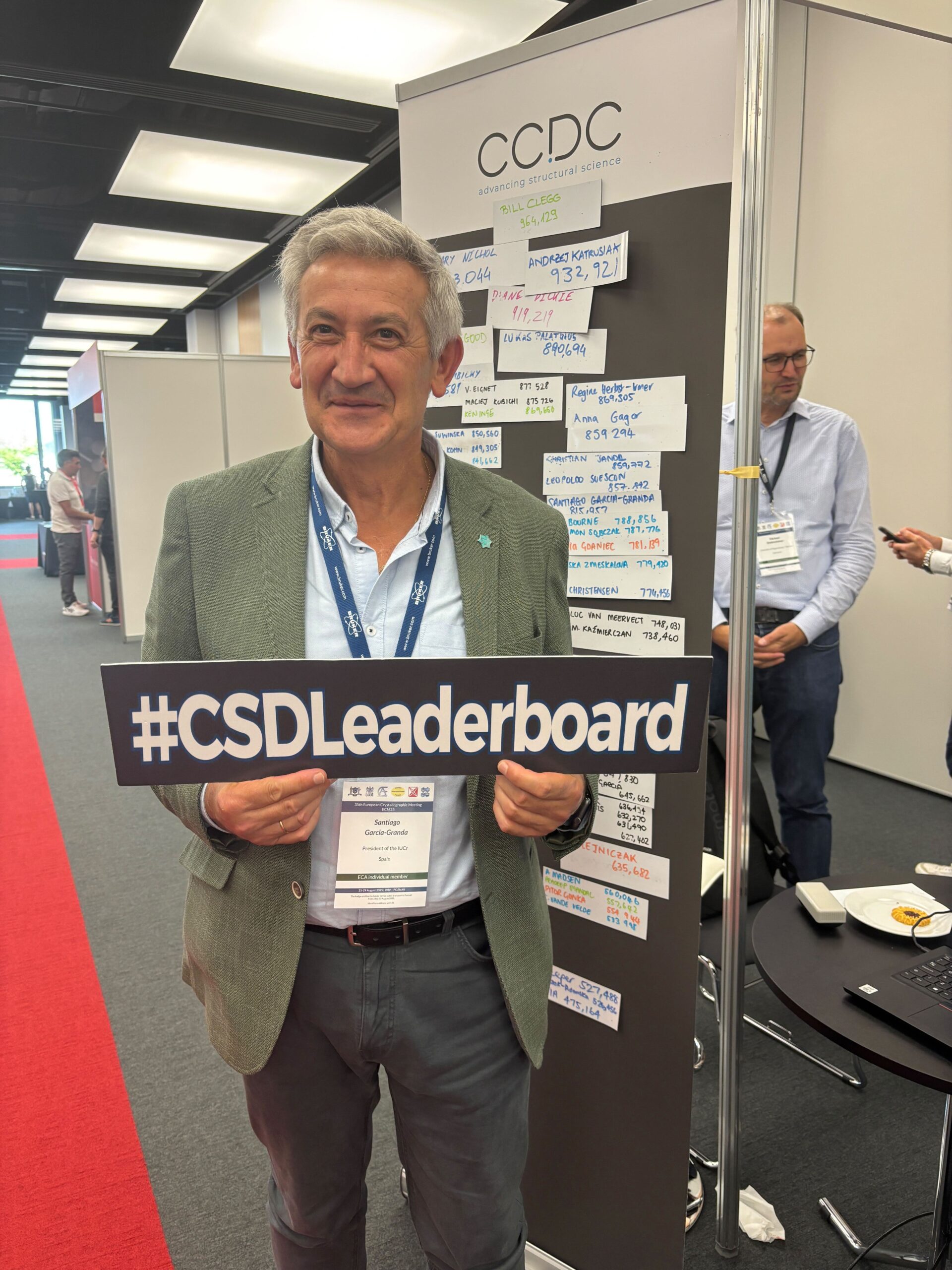 A man in a conference room, holding a sign, stands by a board with stickers on it.