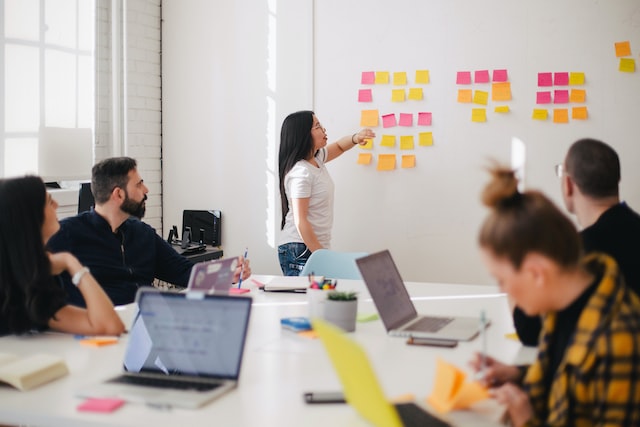 Standing up women pointing to a white board with post-it notes and four people listening and taking notes.