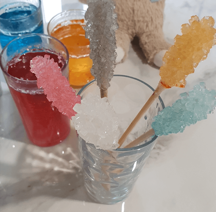 Glasses of different coloured liquid behind a glass containing 5 sticks of sugar crystals in different colours.
